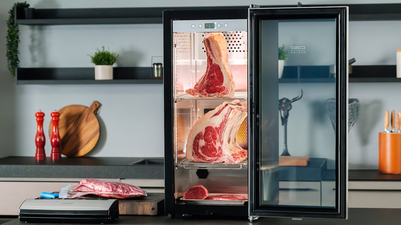 Dry aging fridge with open door, meat cuts, kitchen utensils, shelf, plants.