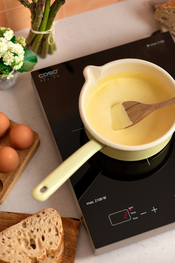 Kochtopf mit Sauce auf Herdplatte, Eier, Brot und Spargel. Kochen in der Küche.