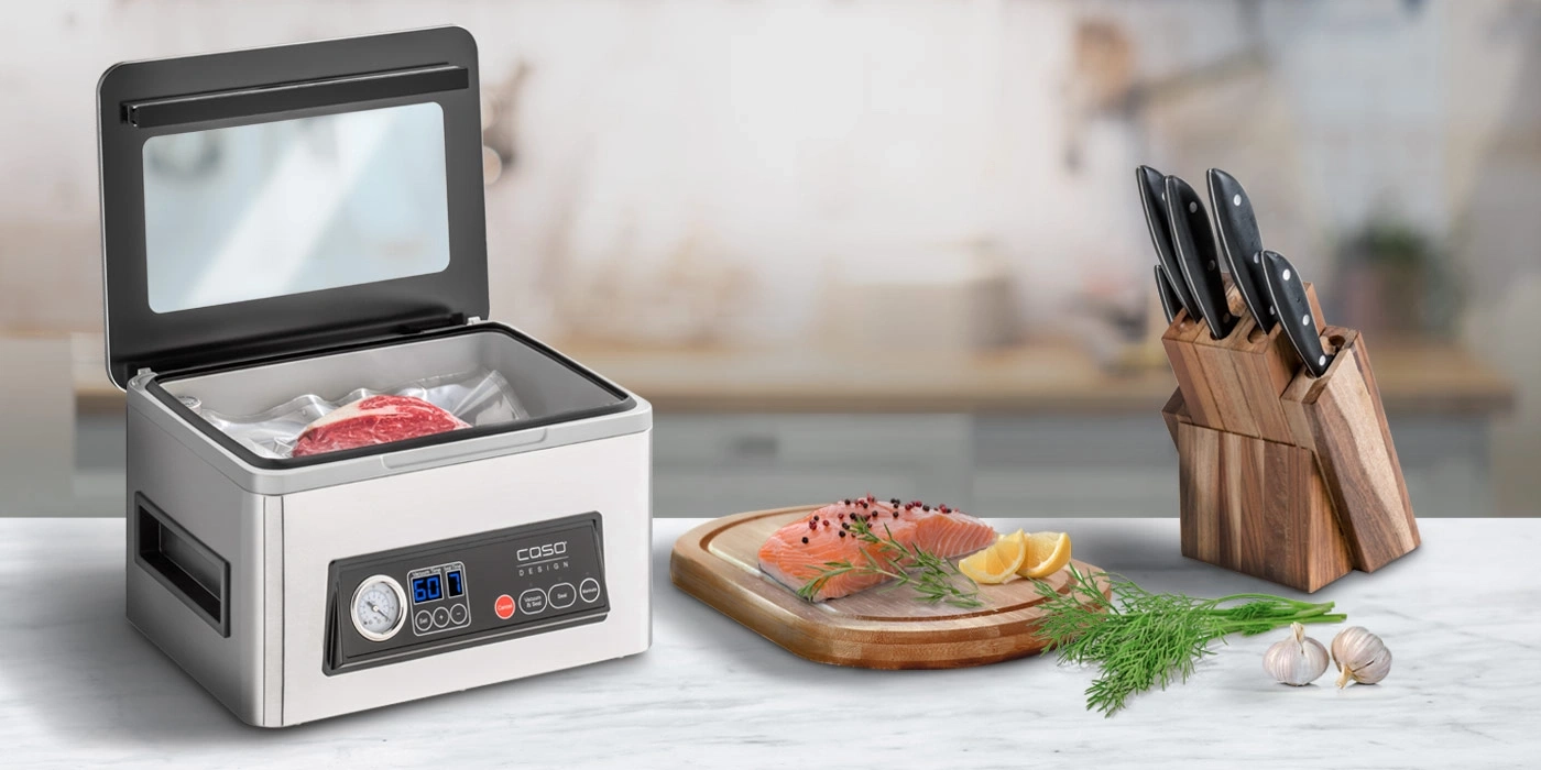 Vakuumiergerät mit Fleisch, Lachs, Messerblock und Knoblauch auf einer Arbeitsplatte.