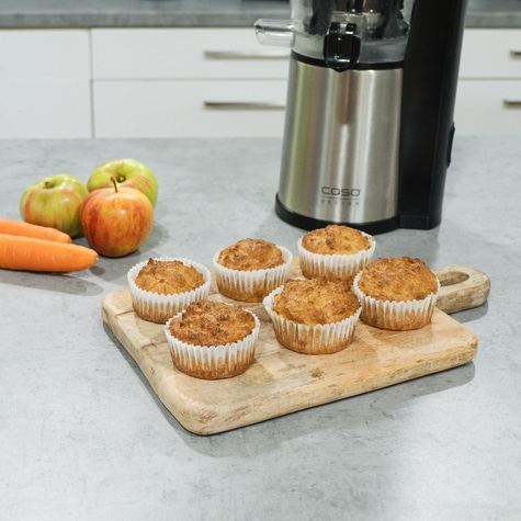 Muffins, Äpfel, Karotten und ein Entsafter auf einer Holzplatte in der Küche.