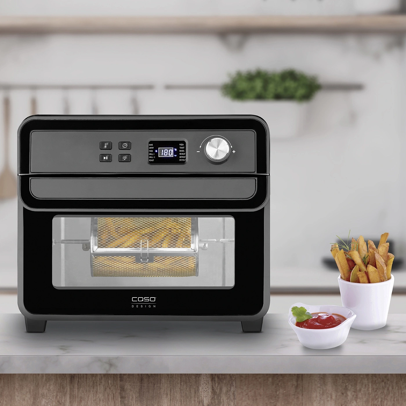 Black air fryer with french fries, ketchup, and herbs on a table.