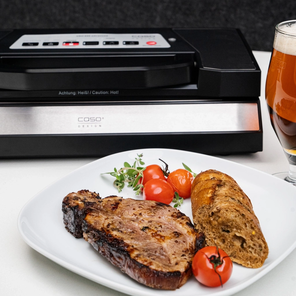 Gebratenes Steak mit Tomaten, Brot und Bier neben Vakuumiergerät.