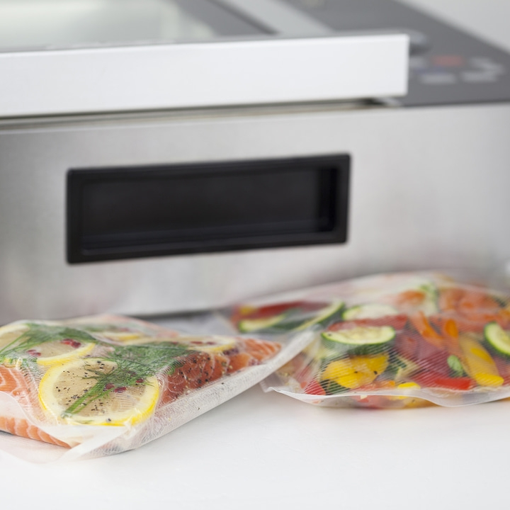 Vakuumiergerät mit Fisch und Gemüse in Beuteln auf weißem Tisch