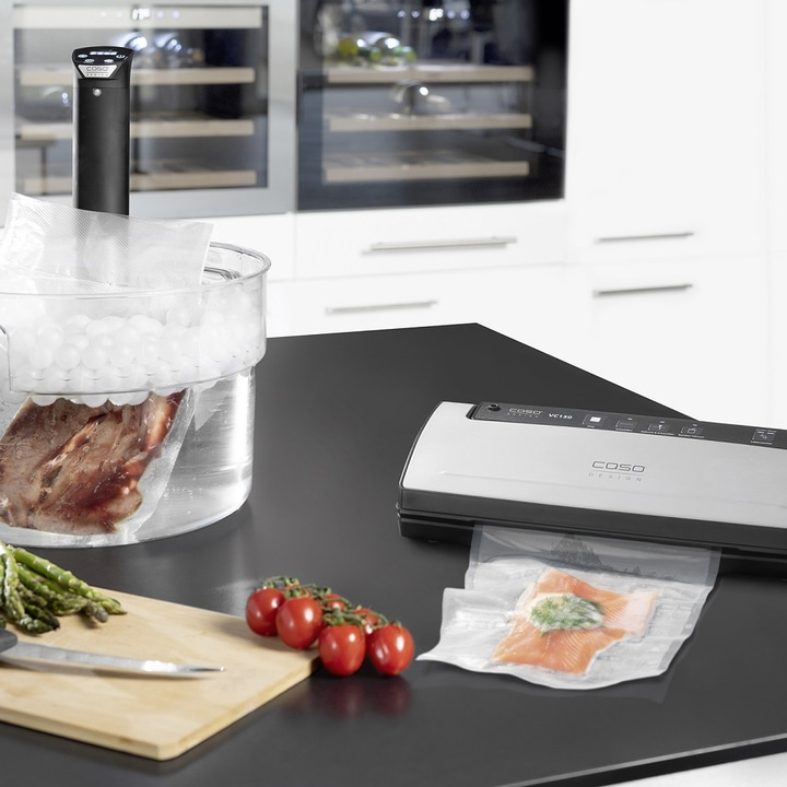 Sous-Vide-Gerät mit Lebensmitteln, Vakuumierer und Gemüse auf schwarzem Tisch.