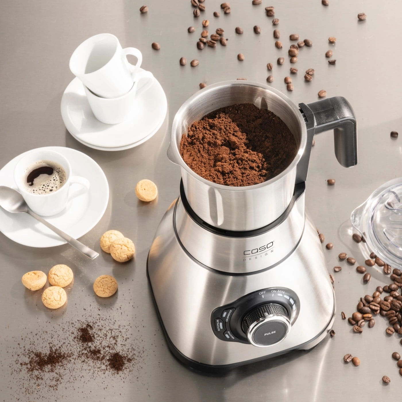 Coffee grinder with coffee, cups, and cookies on a shiny surface.