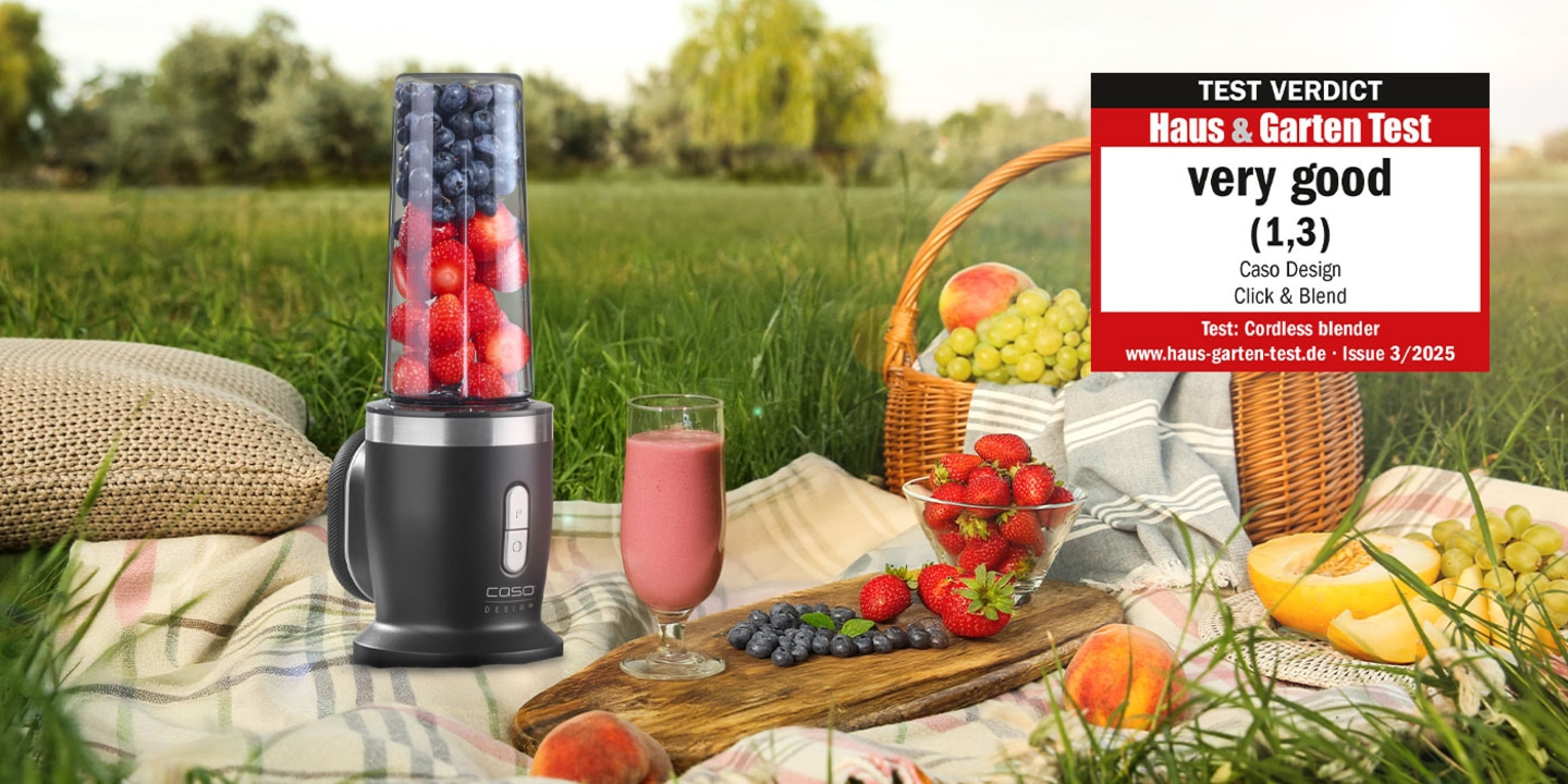 A blender with fruits and a glass of smoothie on a picnic blanket outdoors.