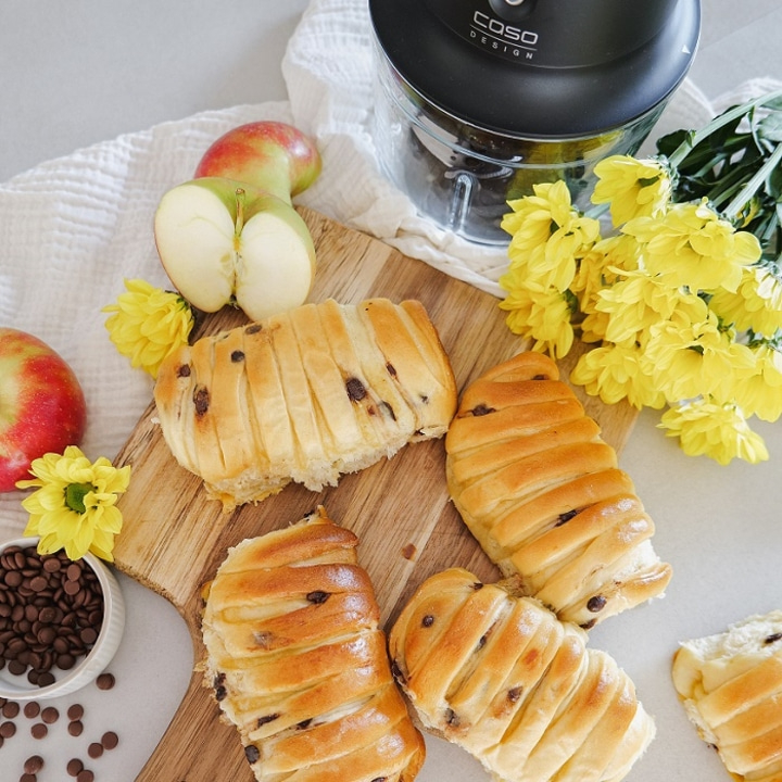 Brot, Äpfel, Schokoladenstückchen und gelbe Blumen auf einem Holzbrett.