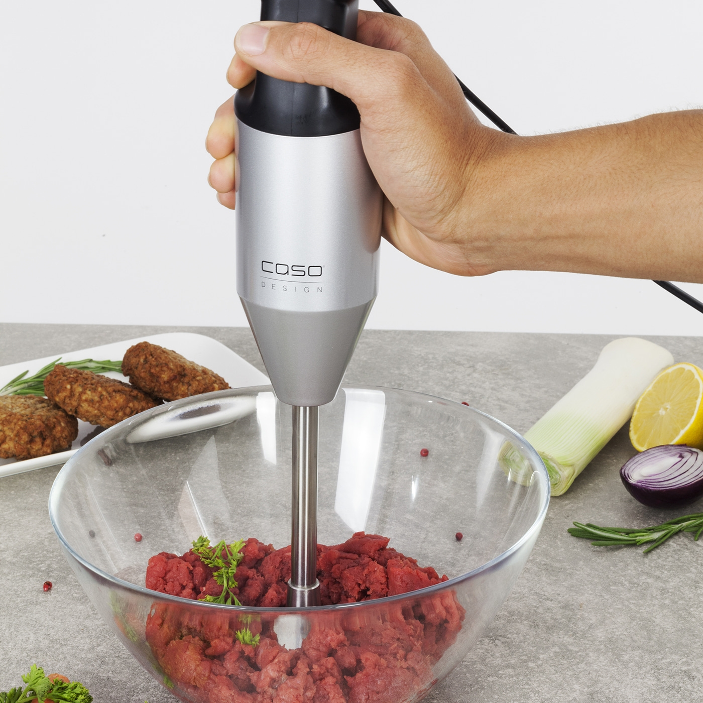 Hand hält Stabmixer über Schüssel mit Hackfleisch, daneben Gemüse und Fleisch