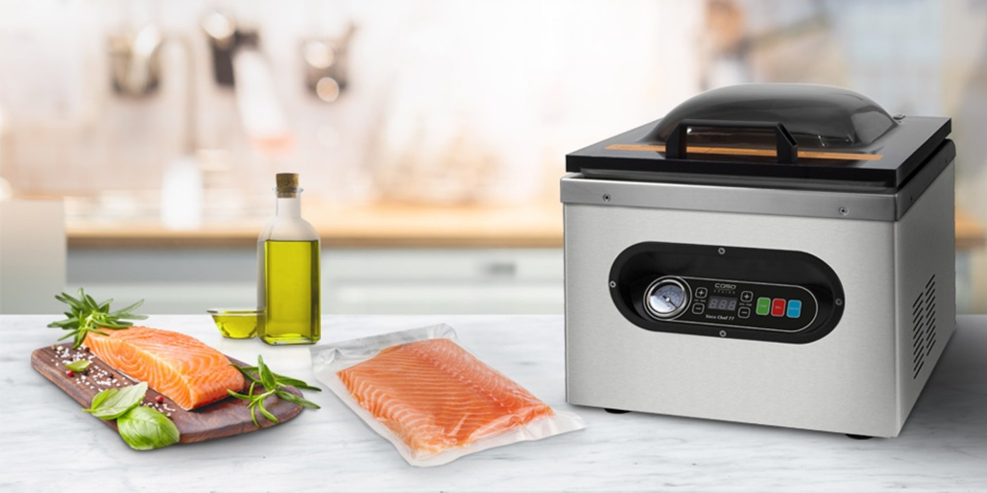 Vacuum sealer with salmon fillet, oil, and herbs on a kitchen table setting.