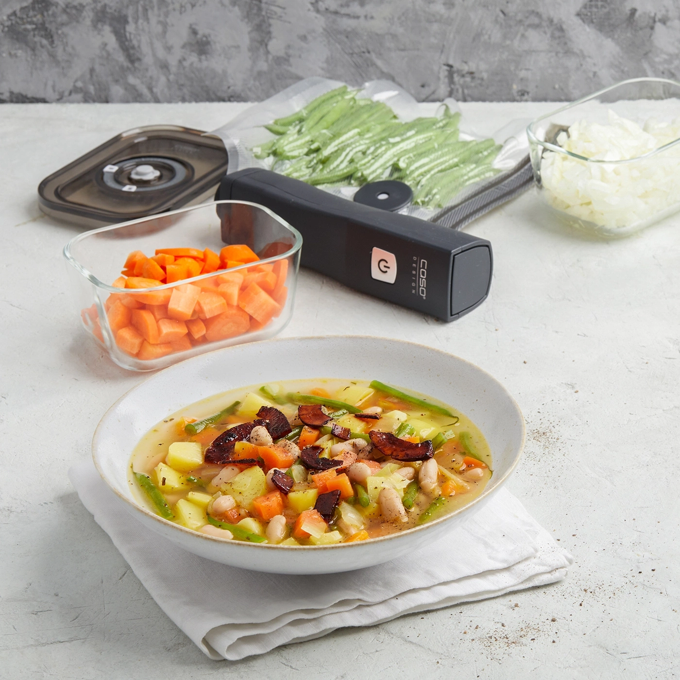 Gemüsesuppe mit Zutaten, Vakuumierer und vorbereitetem Gemüse in Behältern.