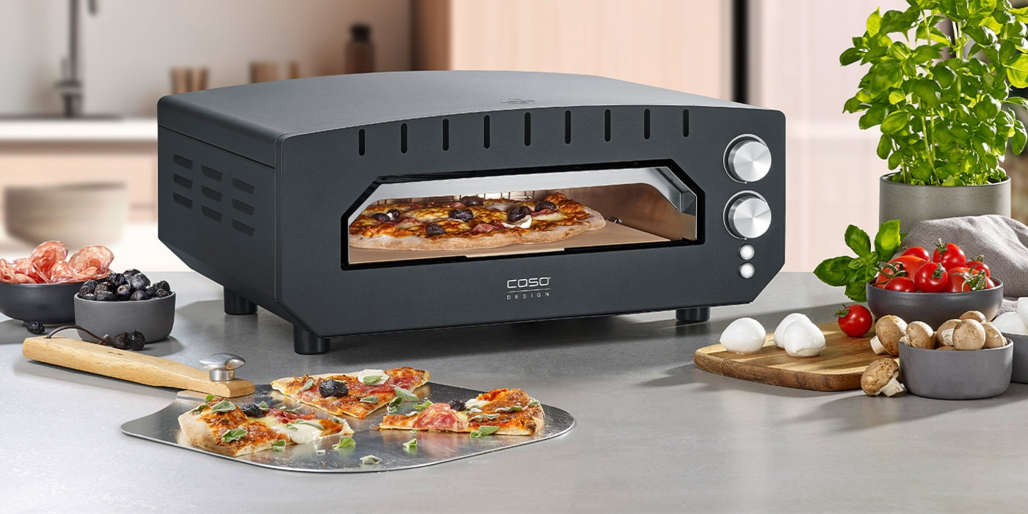 Pizza oven with pizza, ingredients, and pizza slices on a countertop.