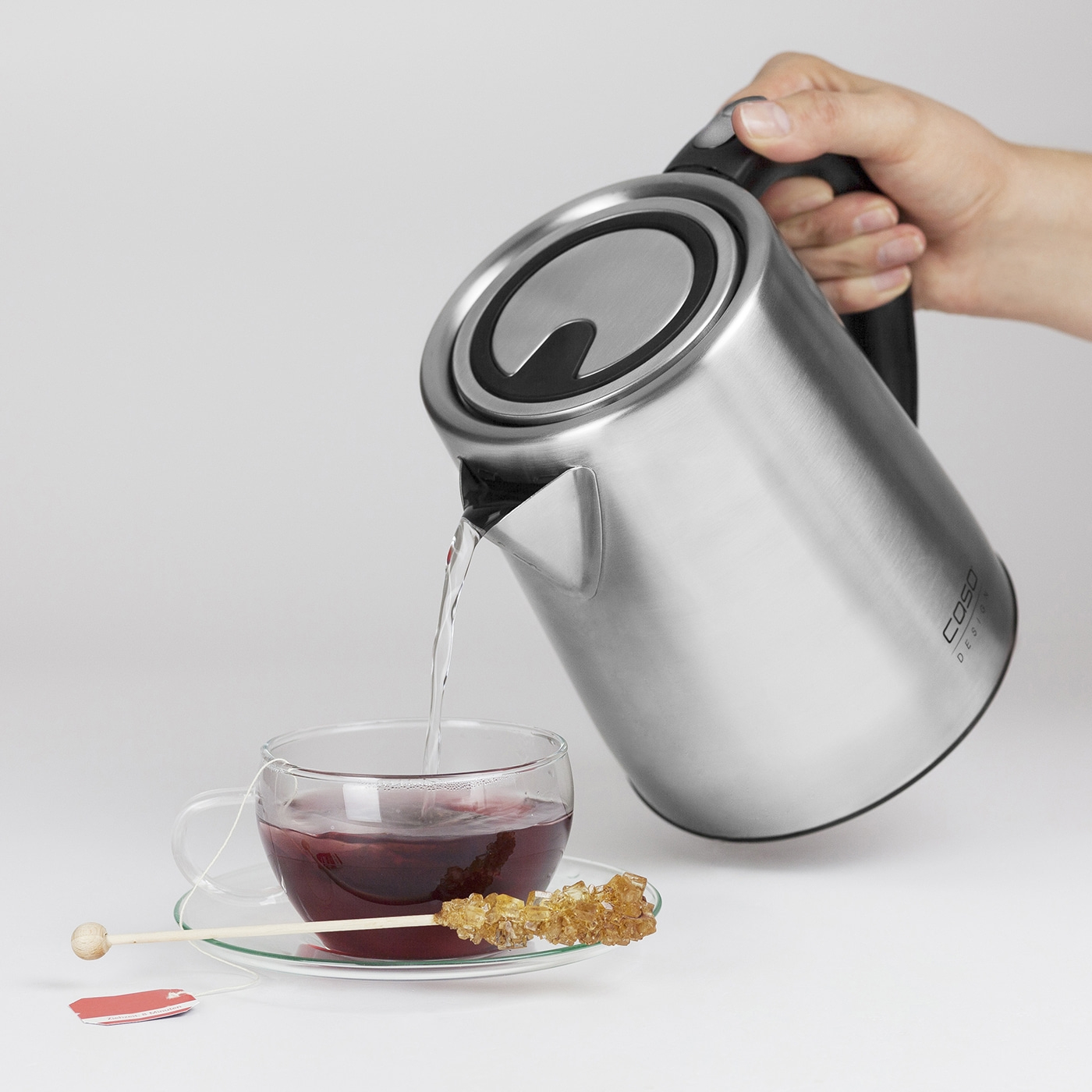 Eine Person gießt Tee aus einem Wasserkocher in eine Tasse mit Kandiszucker.
