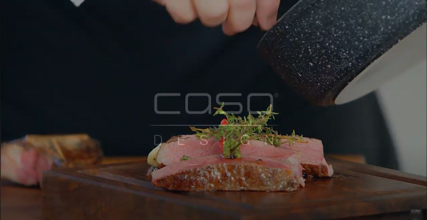 Ein Koch serviert ein Steak mit Kräutern auf einem Holzbrett.