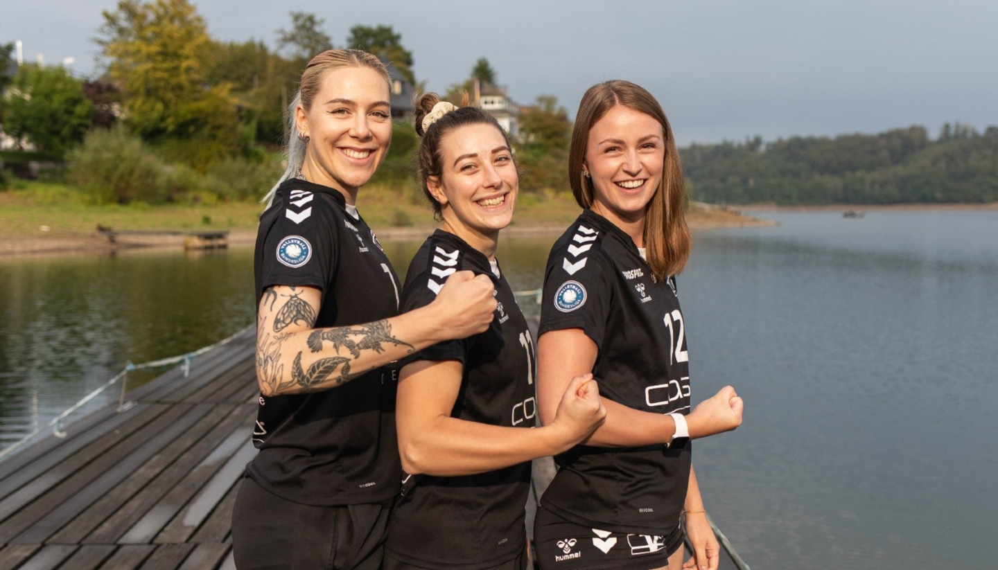 Drei lächelnde Frauen in Sportkleidung posieren am Seeufer.