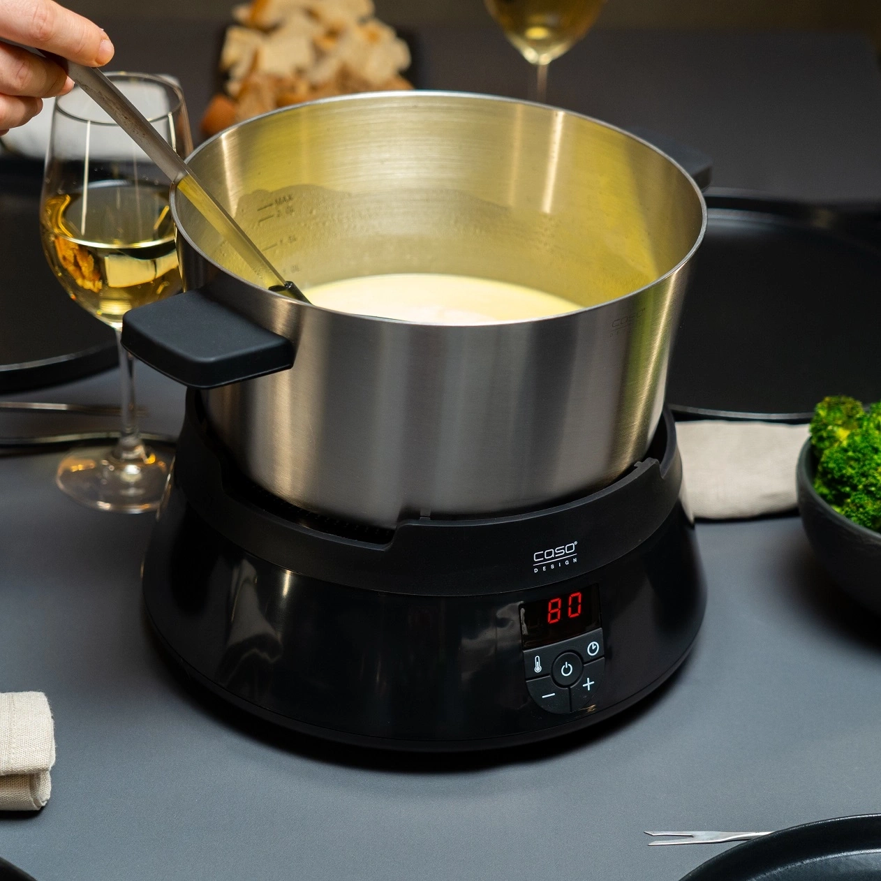 Käsefondue-Set mit Topf, Tisch, Essen, Hand, Glas, Temperaturanzeige.