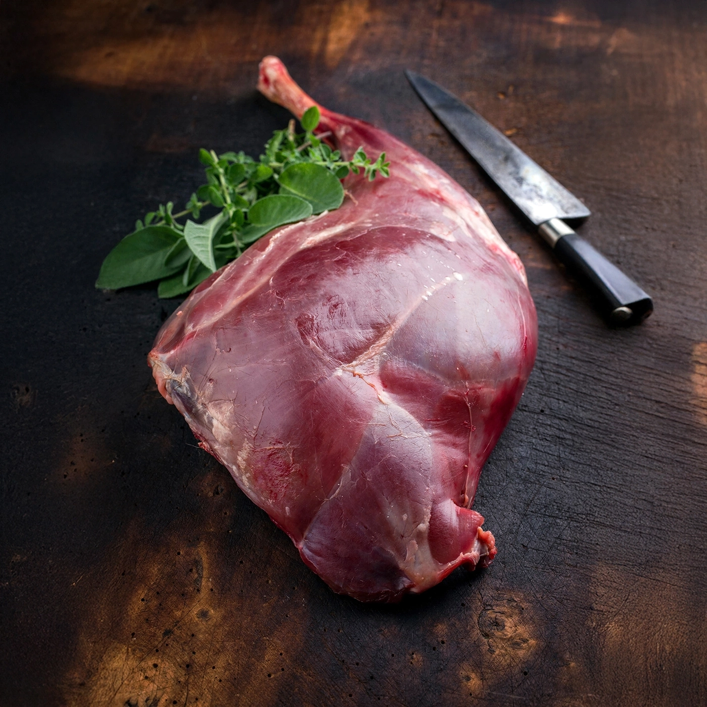 Raw deer leg with herbs and knife on dark wood