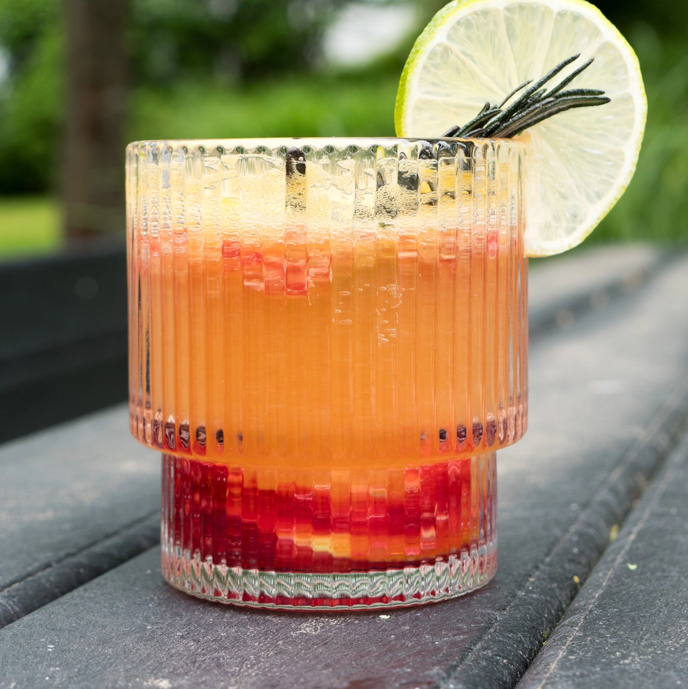 A vibrant red and orange layered drink in a textured glass with a lime slice and rosemary sprig.