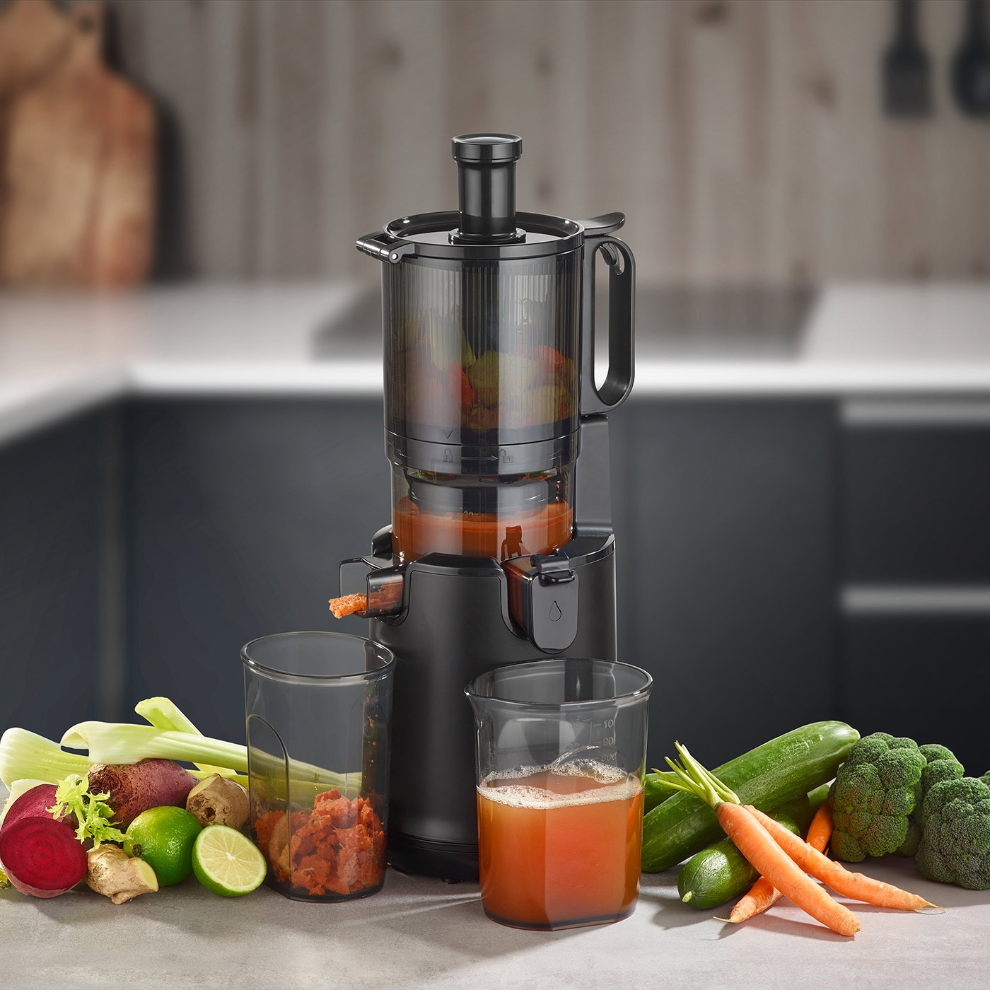 A black juicer with vegetables and juice in glasses on a countertop.