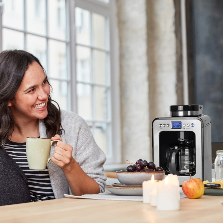 Frau trinkt Kaffee, lächelt, Kaffeemaschine, Obst, Kerzen, Tisch, Fenster, hell