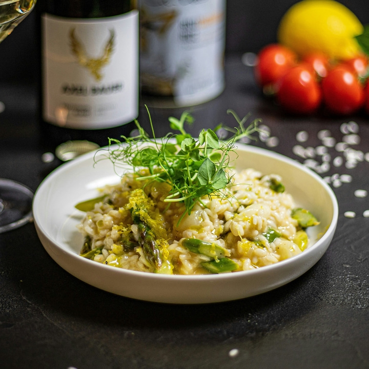 A plate of risotto with asparagus, lemon, and herbs, with wine in the background.