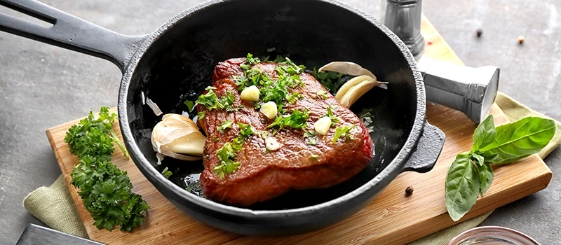 Gebratenes Steak in einer Pfanne mit Knoblauch und Kräutern auf einem Holzbrett.