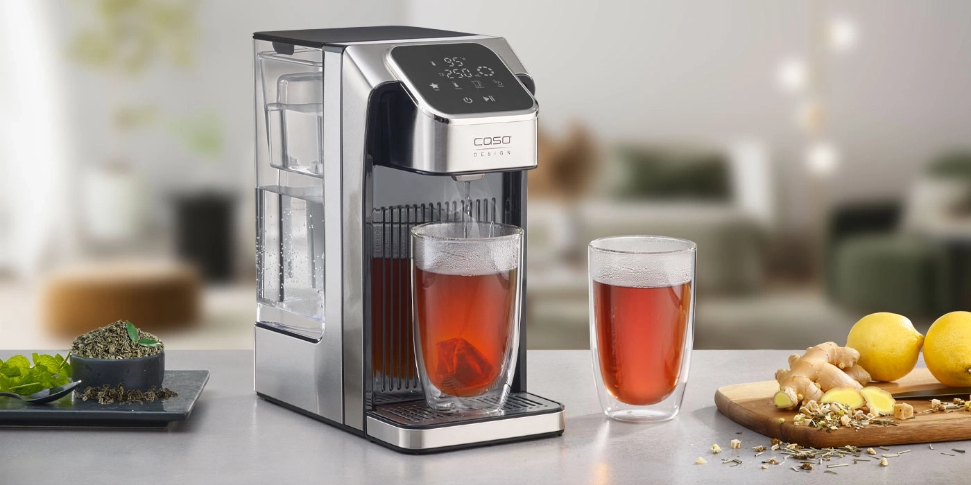 Teemaschine mit zwei Gläsern Tee, Zitrone, Ingwer und Kräutern auf dem Tisch.