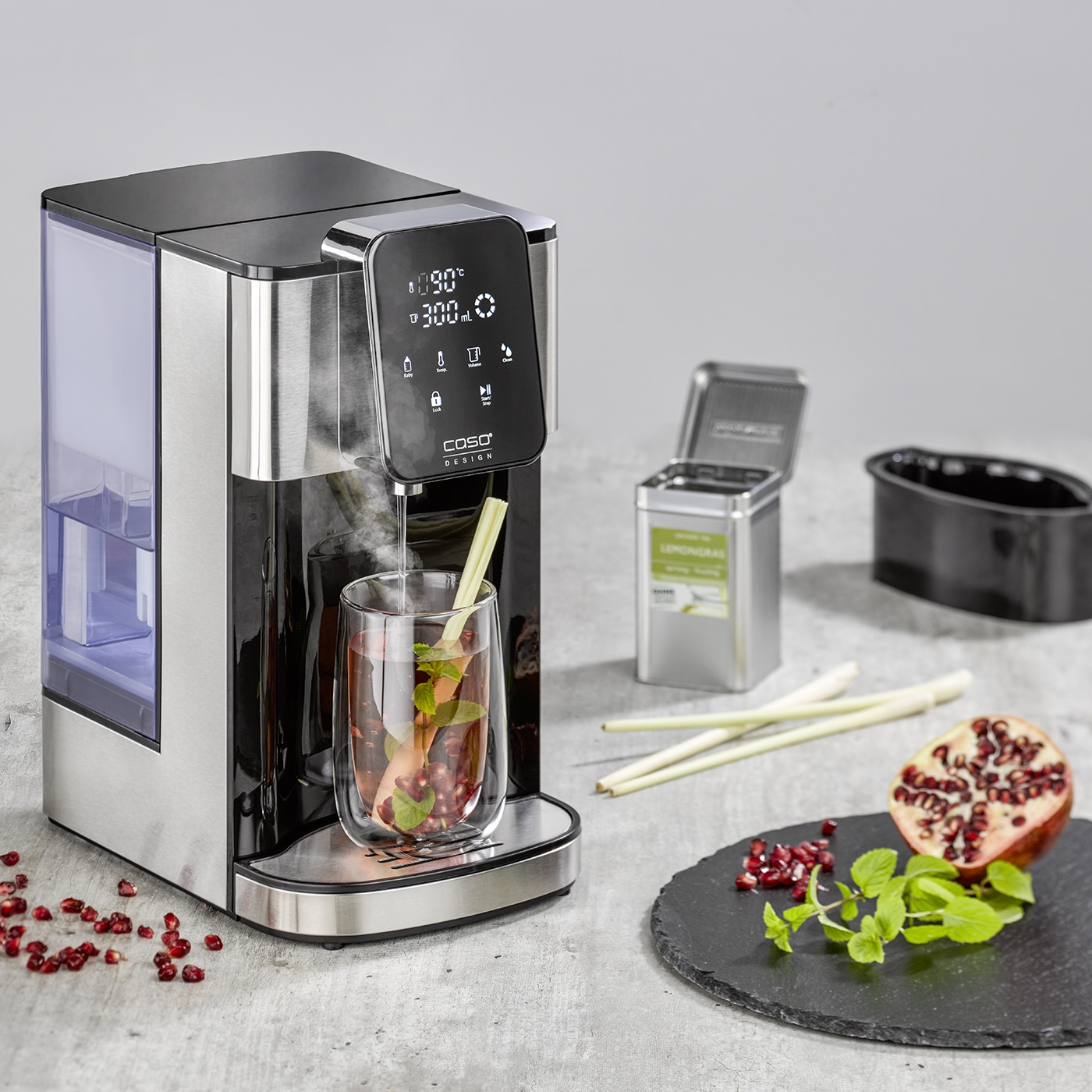 Teemaschine mit Glas, Granatapfel, Minze und Lemongras auf Schieferplatte.