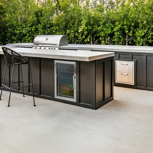 Outdoor-Küche mit Grill, Kühlschrank und Barhocker vor grünen Pflanzen.
