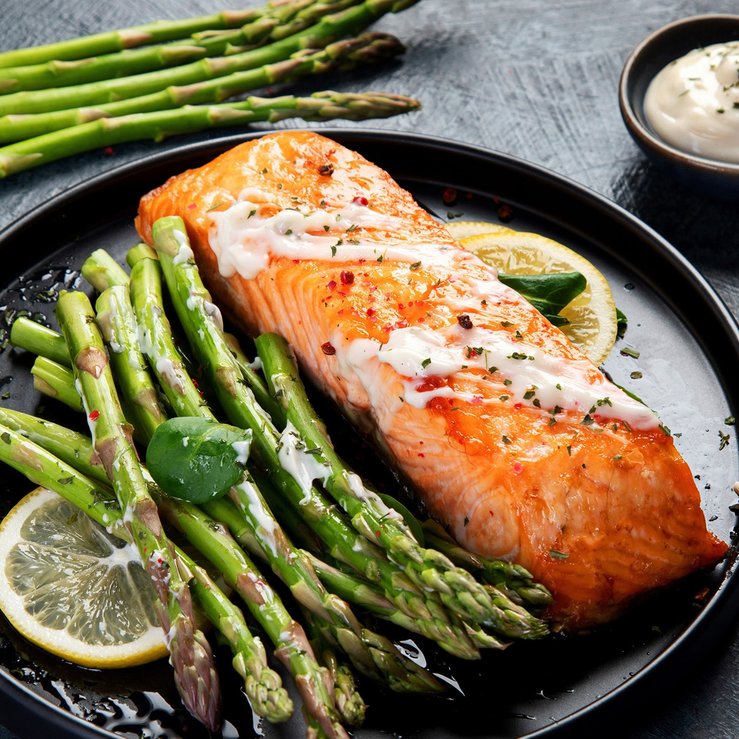 Gebratenes Lachsfilet mit Spargel, Zitrone und Sauce auf einem schwarzen Teller.