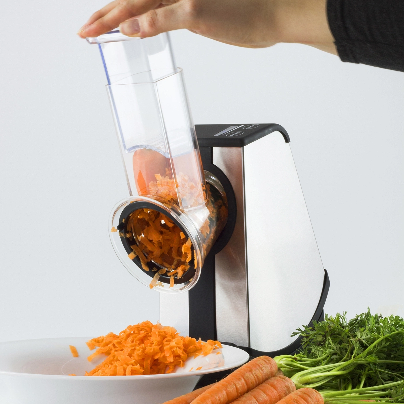 Vegetable slicer with carrots being grated into a bowl, with carrots and greens.