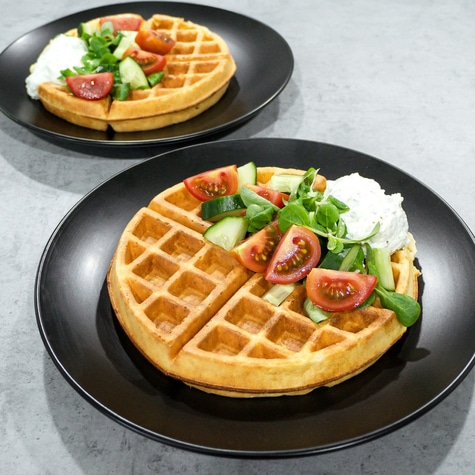 Two waffles with tomatoes, cucumbers, and white cream on black plates.