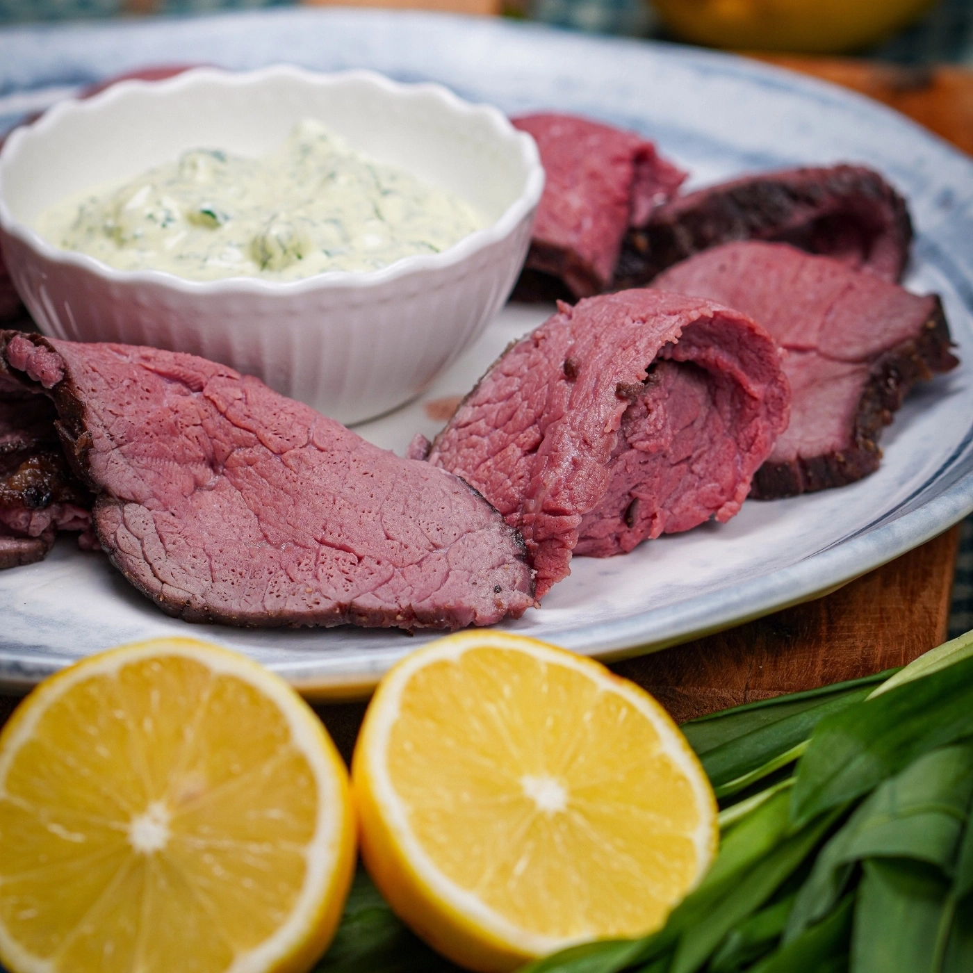 Sliced roast beef with sauce, lemon wedges, and herbs on a plate.
