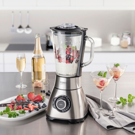 Blender mit Früchten, Getränken und Zutaten auf einer Küchenarbeitsplatte.