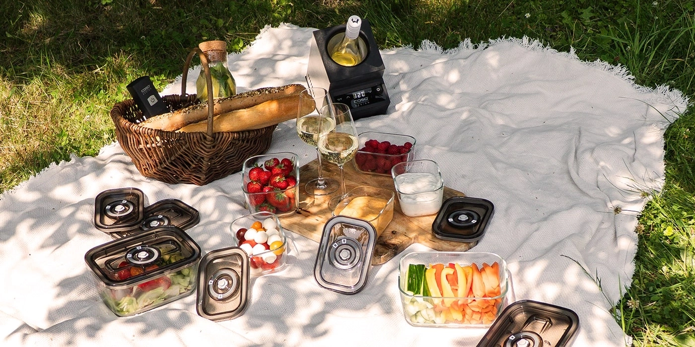 Picknick-Szene mit Essen, Getränken und Decke im Gras an einem sonnigen Tag.