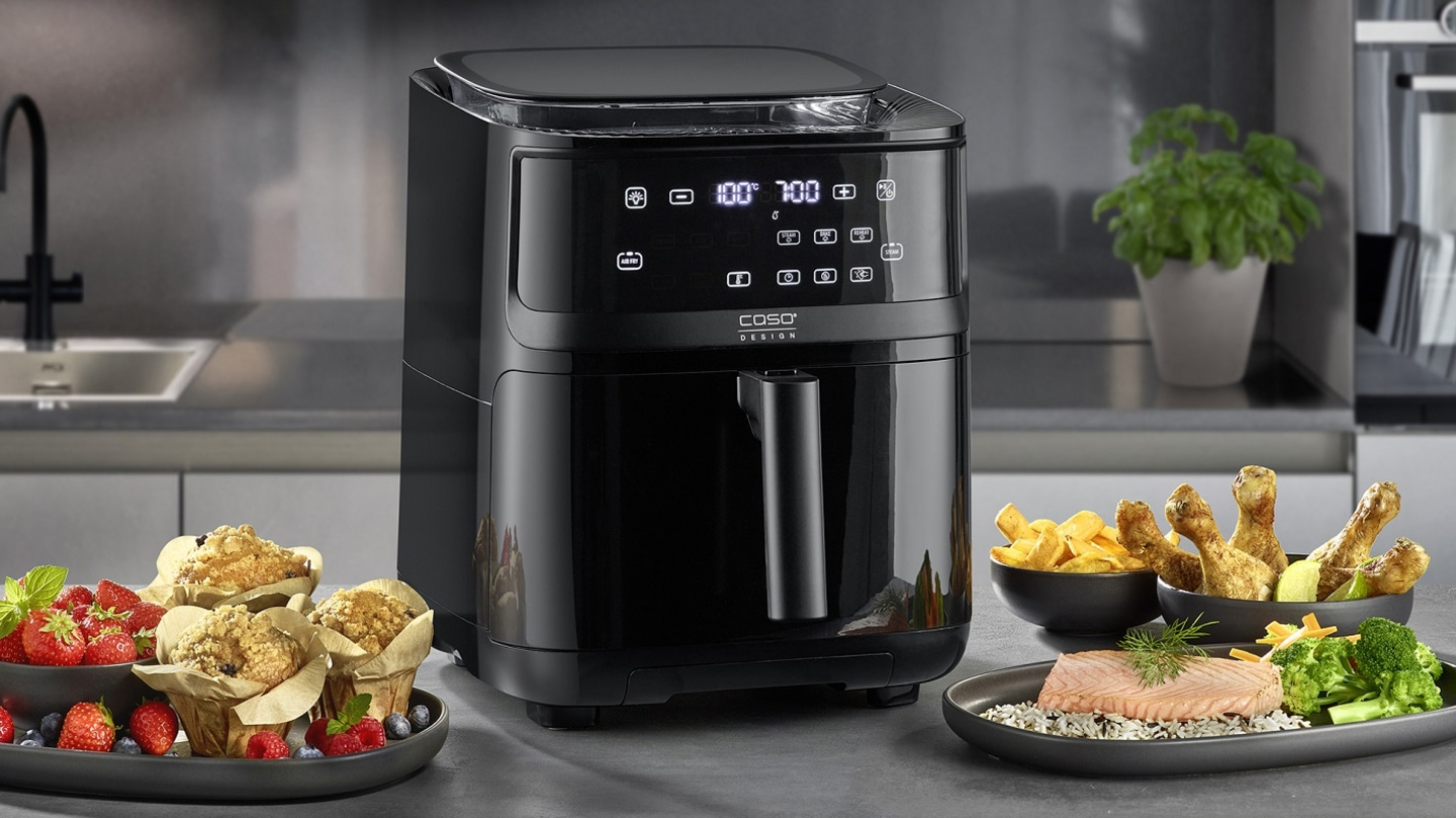 Schwarze Heißluftfritteuse mit Essen, Muffins, Hähnchenschenkeln und Gemüse auf dem Tisch.