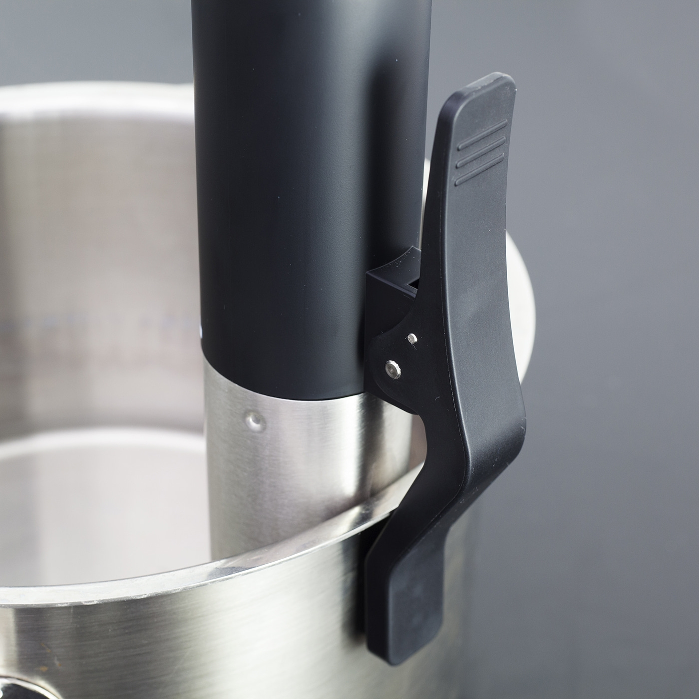 Black device with clamp on metal pot, close-up shot, gray background.