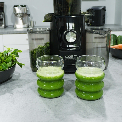 Two green drinks in glasses next to a juicer and ingredients on a counter.