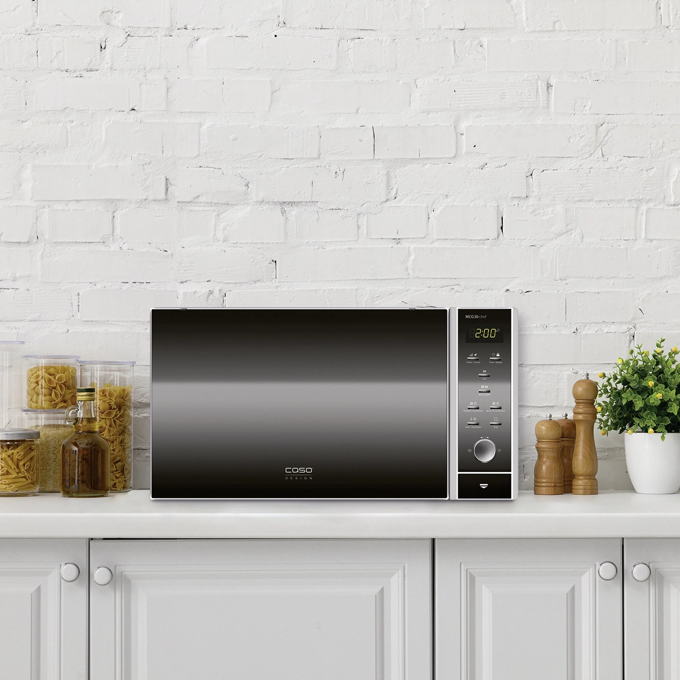Microwave on white cabinet with accessories and potted plant in front of white brick wall.