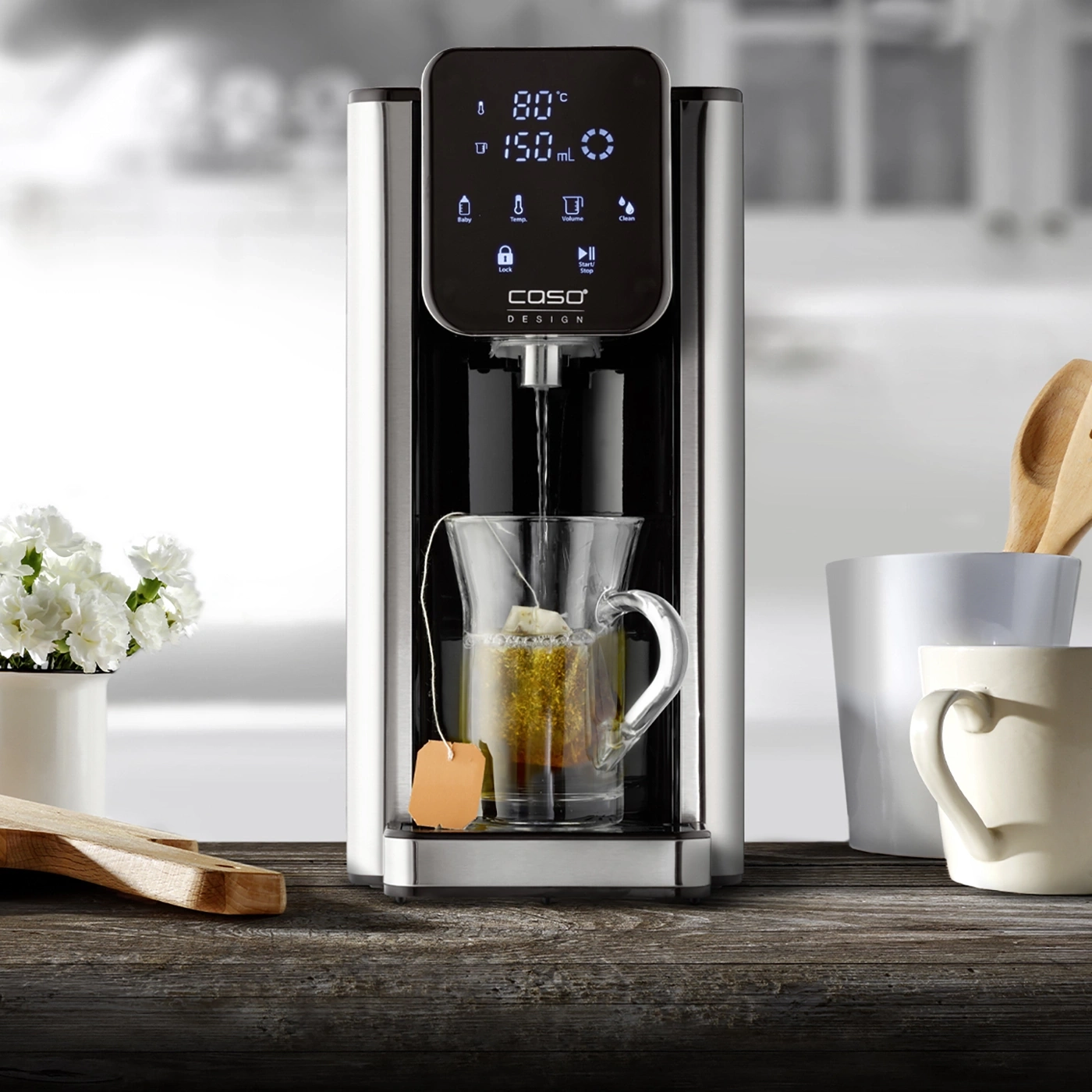 Tea machine with glass cup, tea bag, wooden board and flowers on wooden table
