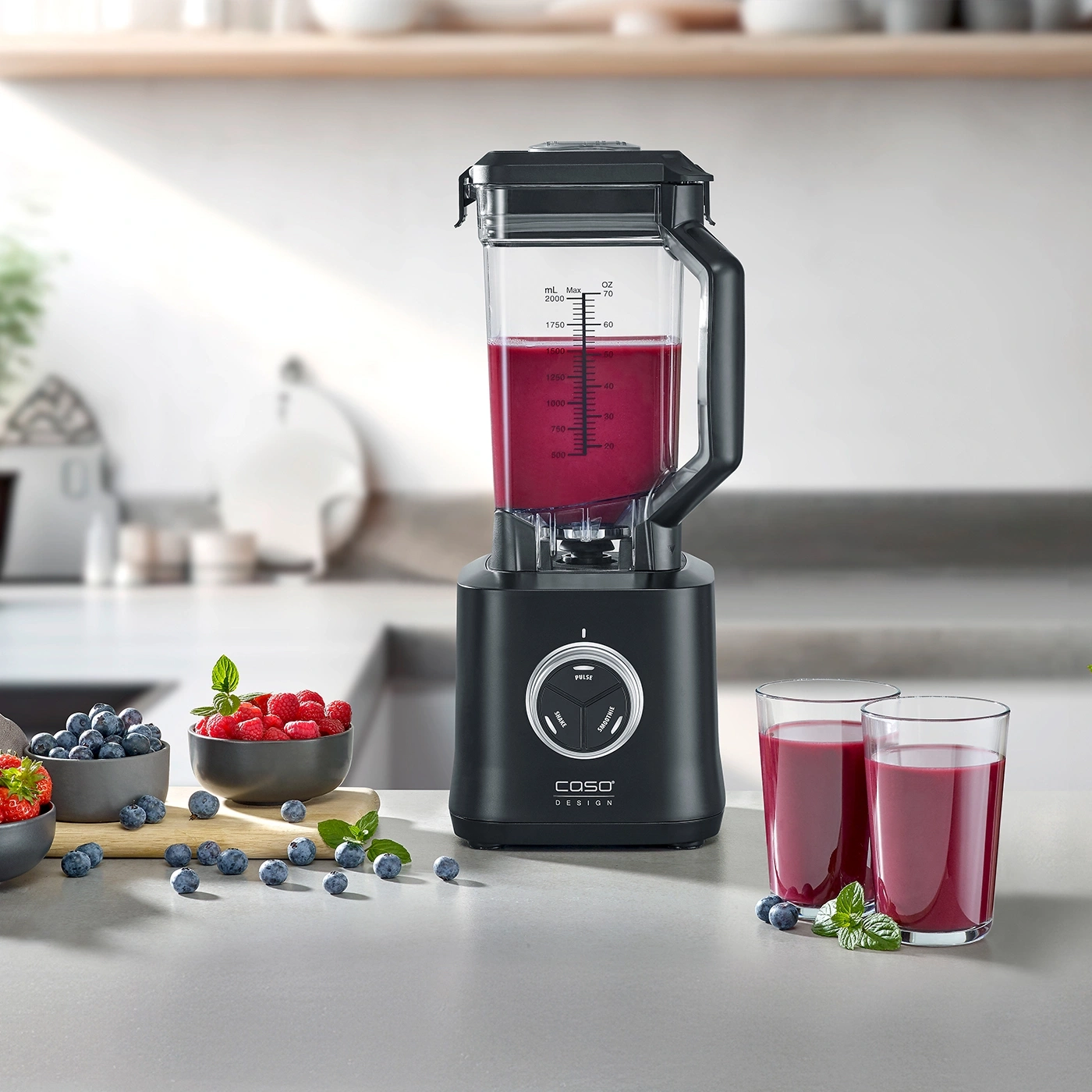 Black blender with smoothie, berries, and glasses on a kitchen countertop.