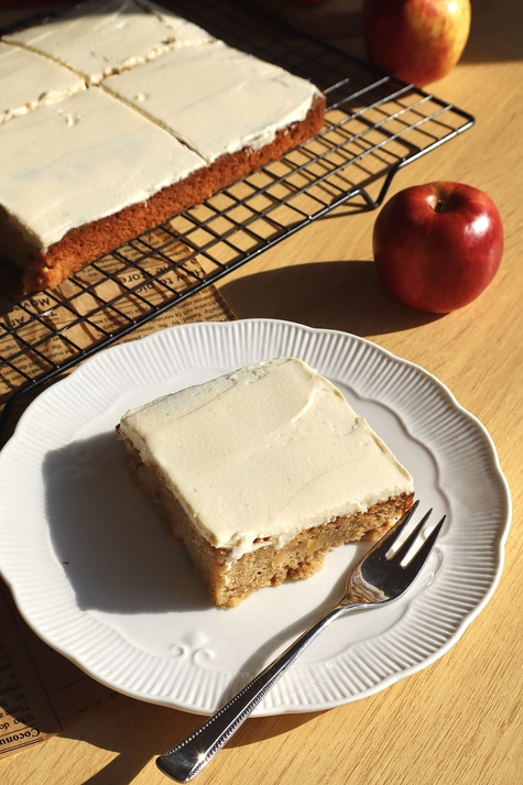 Cake slice on plate with fork, apple, and cake on rack, wooden surface