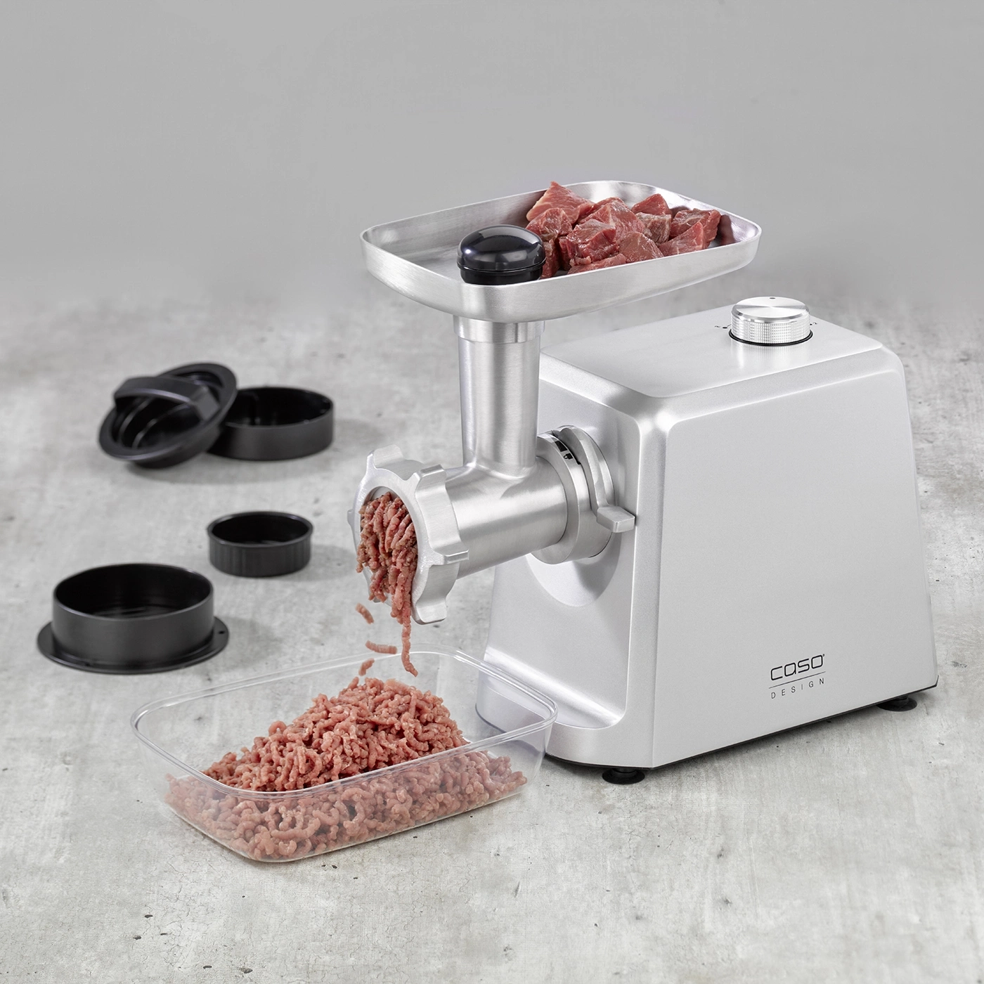Fleischwolf mit Fleisch und Zubehör auf grauem Untergrund. Caso Design.