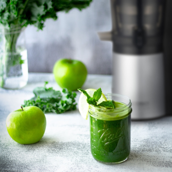 Green smoothie with apples, lime, and mint in a glass, with a juicer nearby.