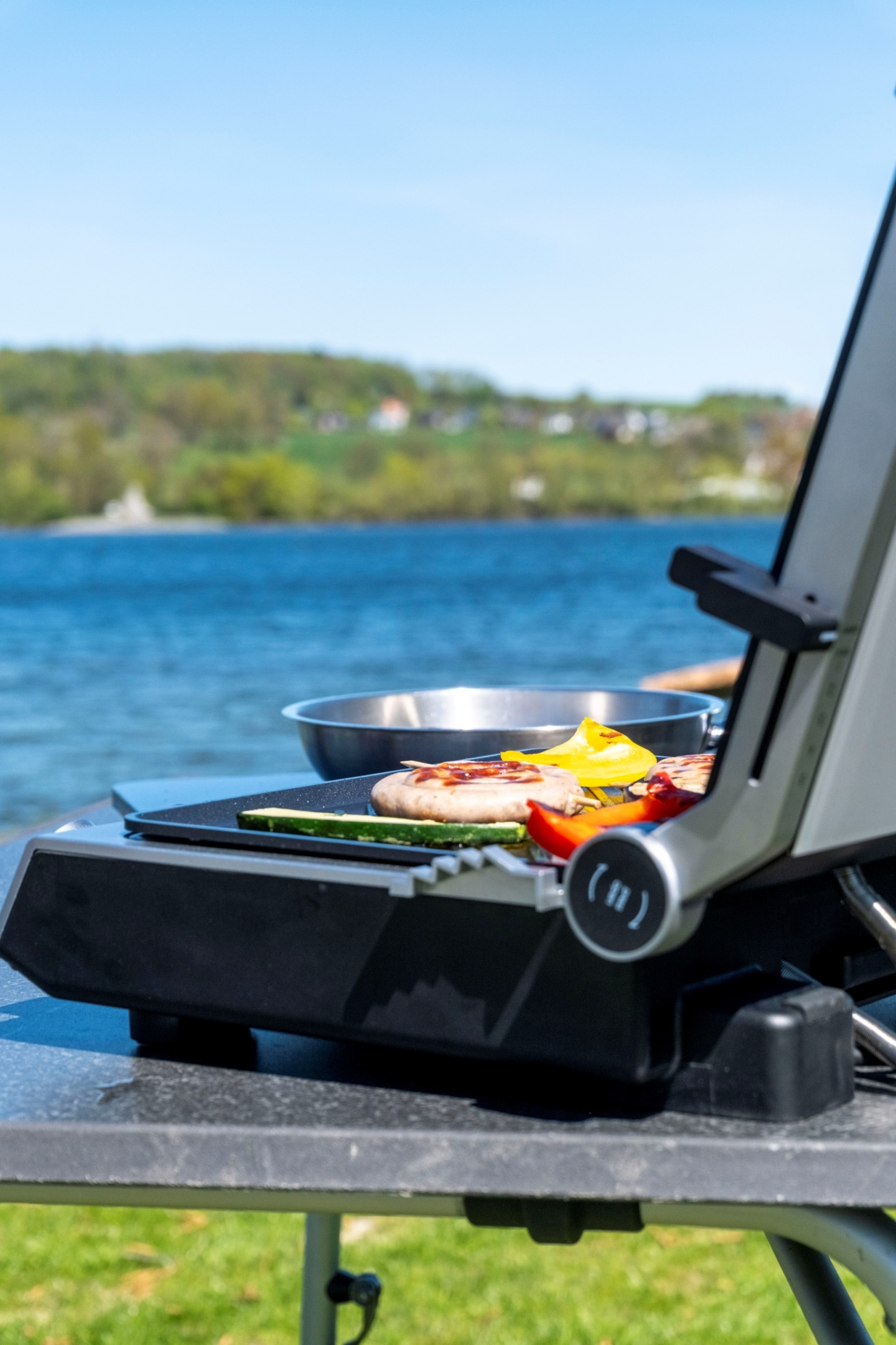 Grill mit Würstchen und Gemüse am Seeufer, blauer Himmel
