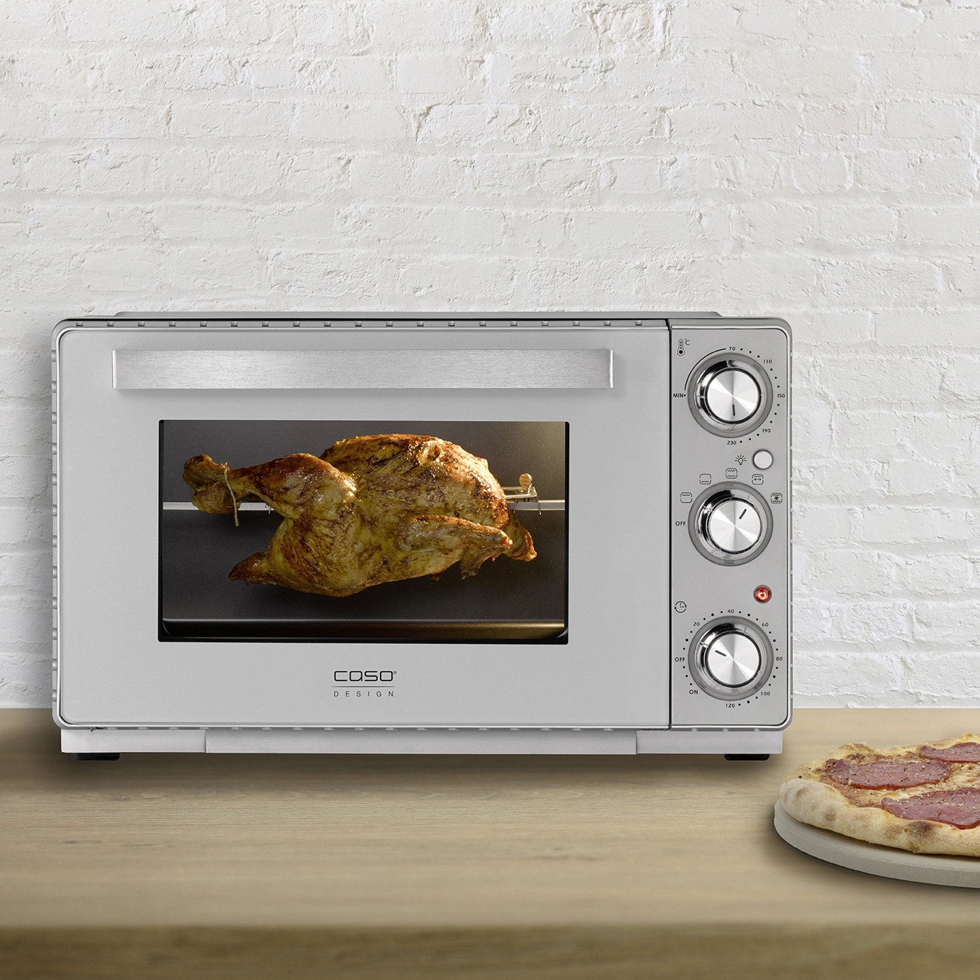 Ofen mit Hähnchen und Pizza auf einem Holztisch vor einer Backsteinmauer