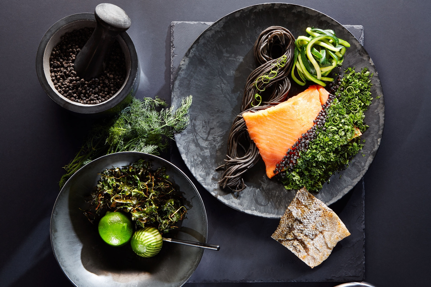 Ein schön arrangiertes Gericht mit Lachs, schwarzen Nudeln, Zucchini und Beilagen auf dunklem Geschirr.