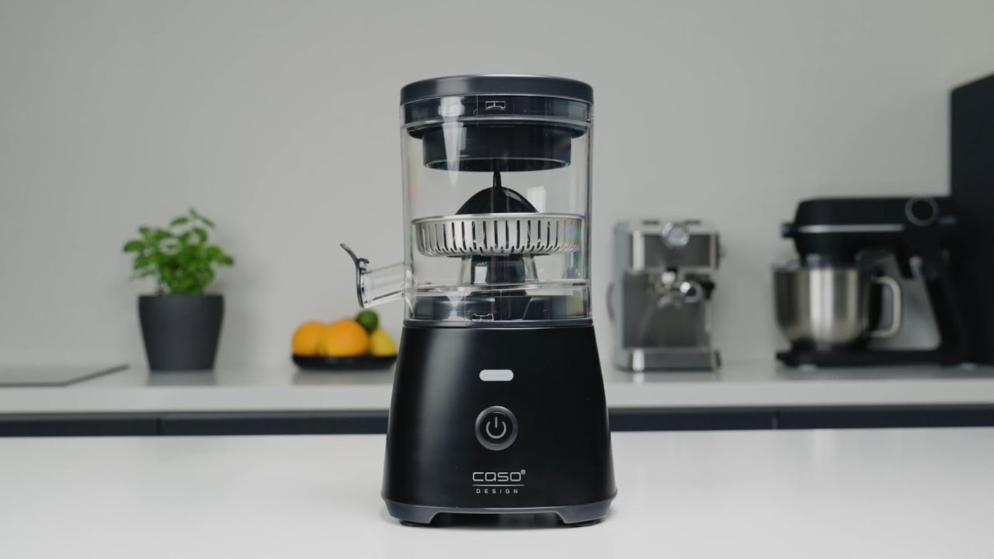 Black juicer on a white surface with other kitchen appliances in the background.
