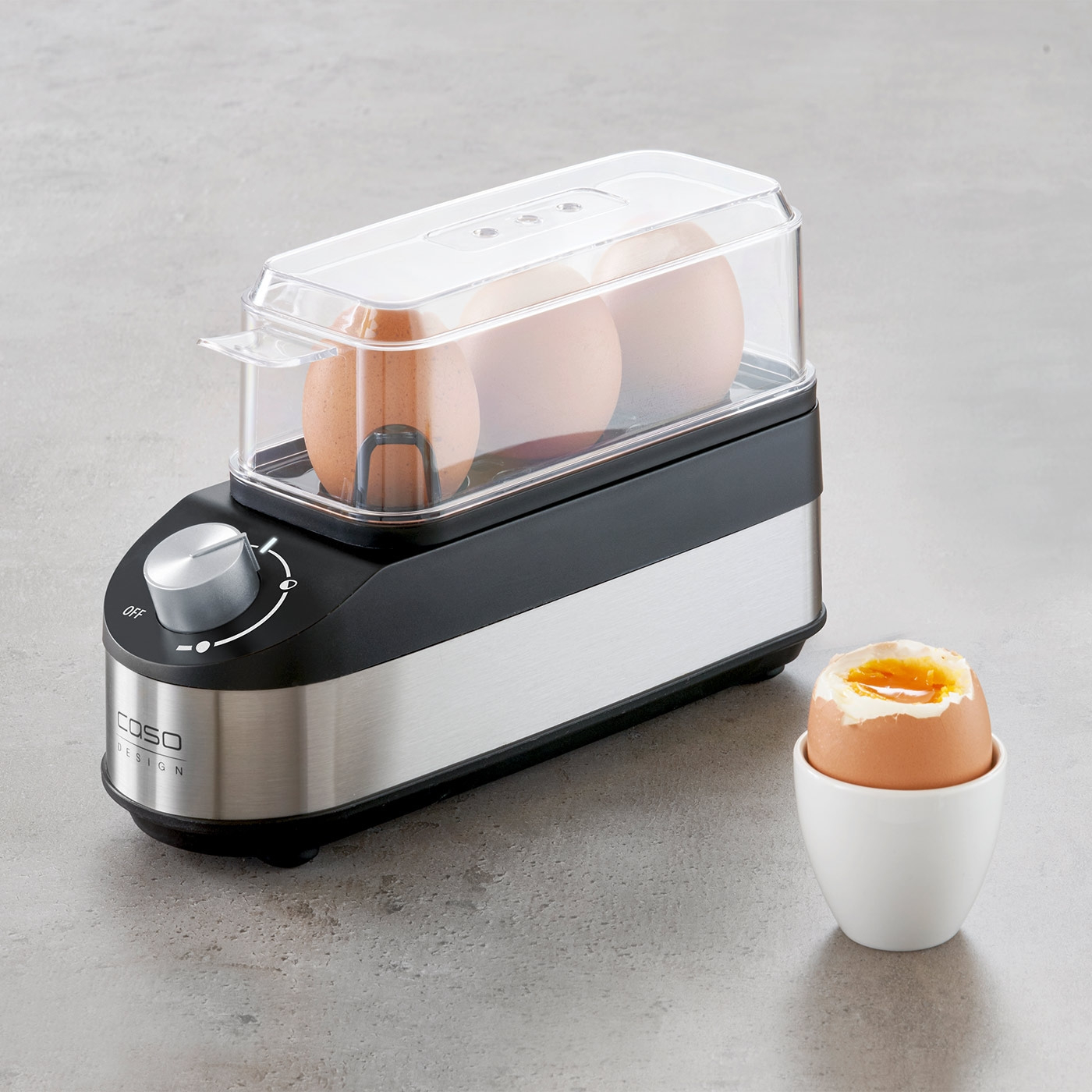 Egg boiler with eggs and a boiled egg in an egg cup on a gray surface.