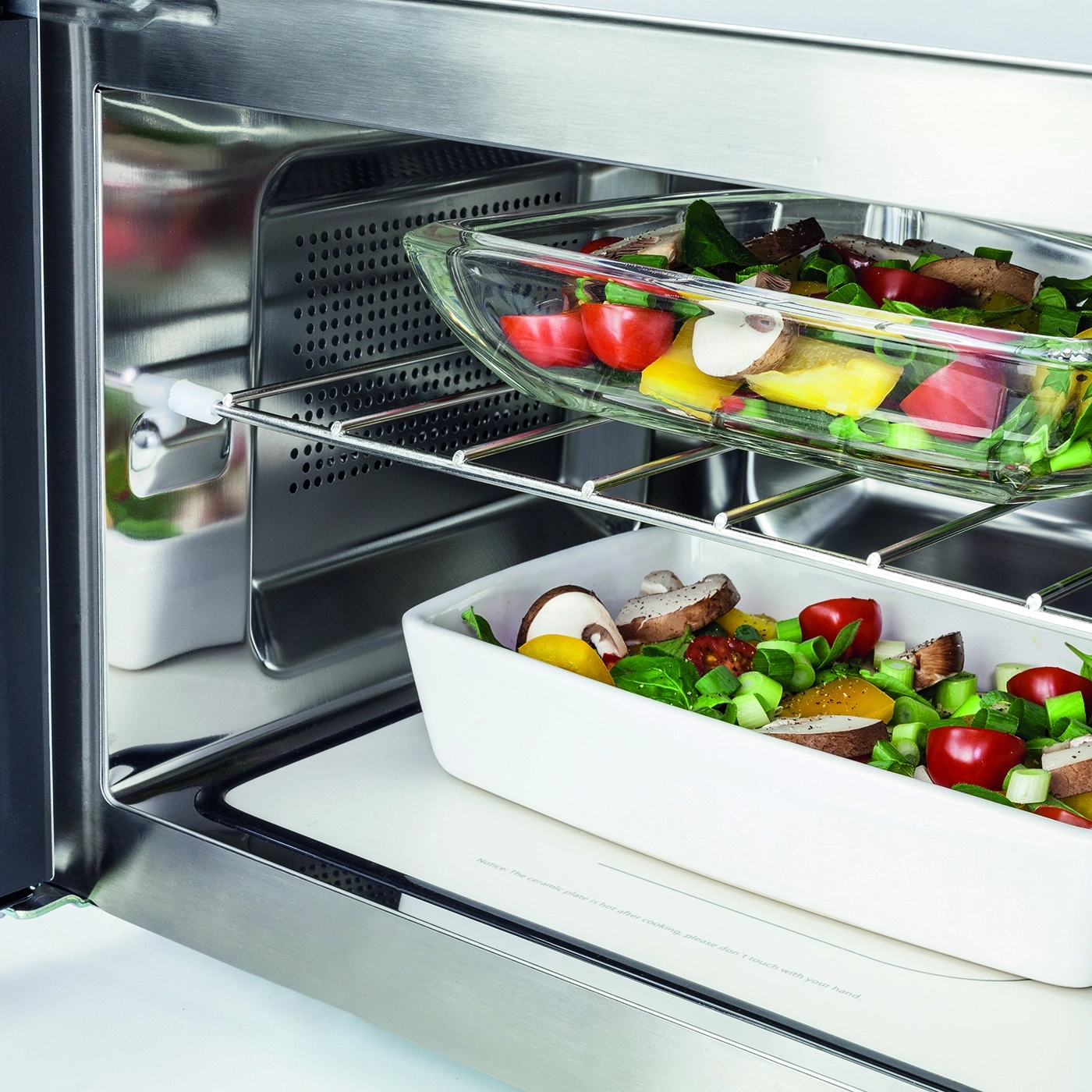 Oven with vegetables in glass and ceramic containers. Stainless steel interior, cooking utensils.