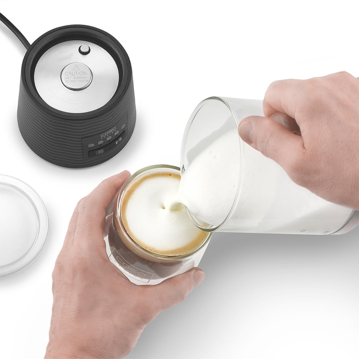 Milch wird in ein Glas mit Kaffee gegossen, daneben ein Milchaufschäumer.